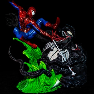 May include: A dynamic figurine depicting Spider-Man in a red and blue suit battling Venom, a black symbiote creature. The scene is set on a vibrant green base with black tendrils, against a black background. The figurine captures a moment of action.