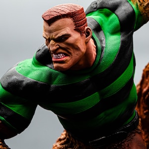 May include: A detailed action figure of a comic book character, Sandman. The figure features a muscular physique, with a green and black striped shirt. The character has a stern expression, with red hair and a textured, sand-like appearance on parts of the body.
