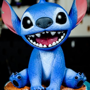 May include: A blue Stitch figurine with large black eyes, a wide grin revealing sharp teeth, and large pink ears. The figurine is seated on a small, round base with a sandy texture.