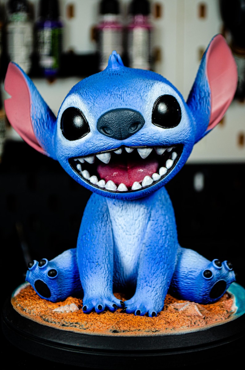 May include: A blue Stitch figurine with large black eyes, a wide grin revealing sharp teeth, and large pink ears. The figurine is seated on a small, round base with a sandy texture.