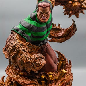 May include: A detailed statue of a comic book character, featuring a muscular figure with a determined expression. The character wears a green and black striped shirt and brown pants. The base is adorned with gold coins and bars, and the figure is surrounded by a textured, brown, sand-like substance.