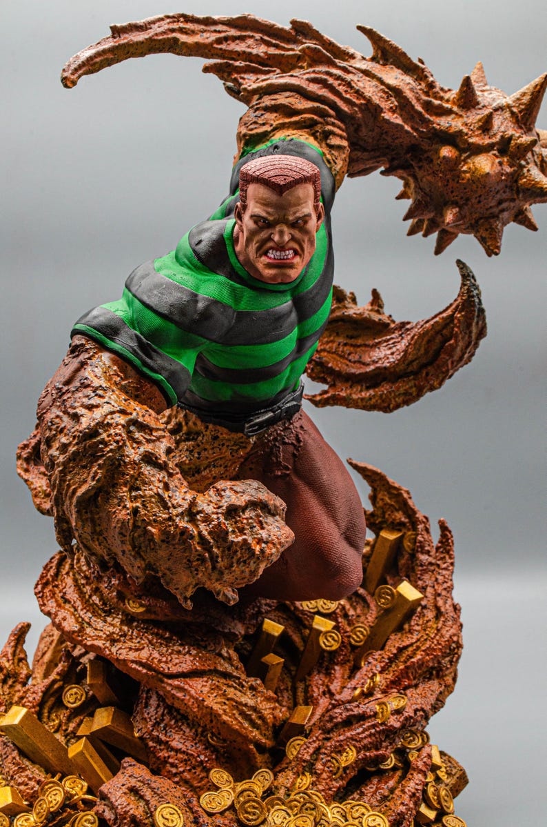 May include: A detailed statue of a comic book character, featuring a muscular figure with a determined expression. The character wears a green and black striped shirt and brown pants. The base is adorned with gold coins and bars, and the figure is surrounded by a textured, brown, sand-like substance.