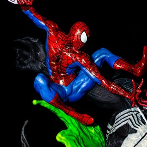 May include: A dynamic figurine of Spider-Man in a classic red and blue suit, poised mid-action with a web strand. The figure is set against a black background, with green and black elements suggesting a battle scene. The figurine is a collectible item.