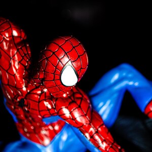 May include: A detailed figurine of Spider-Man, featuring a red and blue costume. The figure's head is red with a white eye, and the costume has a web-like pattern. The figure is posed against a dark background.