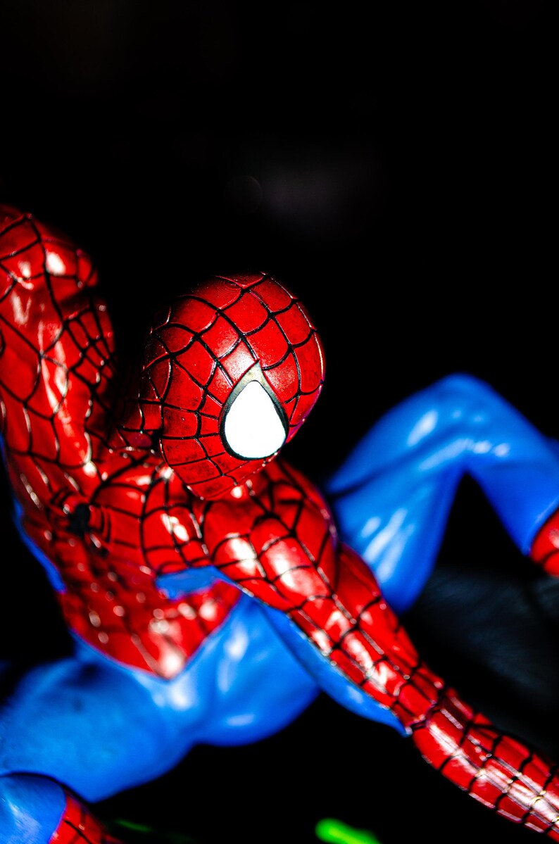 May include: A detailed figurine of Spider-Man, featuring a red and blue costume. The figure's head is red with a white eye, and the costume has a web-like pattern. The figure is posed against a dark background.