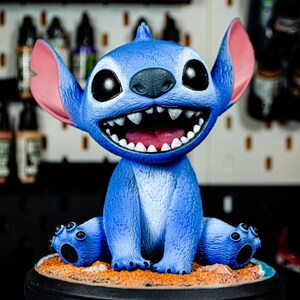 May include: A blue Stitch figurine with large black eyes, pink ears, and a wide, toothy grin. The figurine sits on a sandy base with a black circular stand. The background is blurred, suggesting a studio setting.