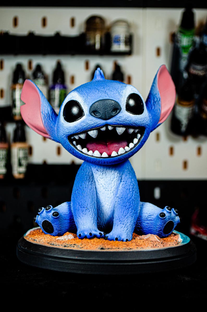 May include: A blue Stitch figurine with large black eyes, pink ears, and a wide, toothy grin. The figurine sits on a sandy base with a black circular stand. The background is blurred, suggesting a studio setting.