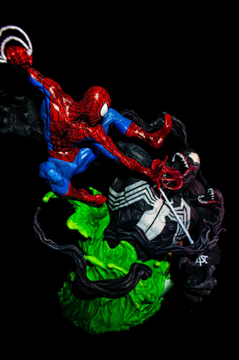 May include: A detailed figurine depicting a battle between Spider-Man and Venom. Spider-Man is in a dynamic pose, clad in a red and blue suit, while Venom, a black symbiote, is in a menacing stance. The base is a vibrant green.