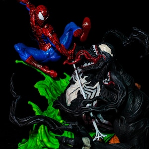 May include: A dynamic action figure scene featuring Spider-Man in a red and blue suit, mid-air, facing off against Venom, a black figure with a white spider symbol. The figures are set against a vibrant green base.