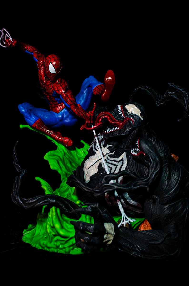 May include: A dynamic action figure scene featuring Spider-Man in a red and blue suit, mid-air, facing off against Venom, a black figure with a white spider symbol. The figures are set against a vibrant green base.