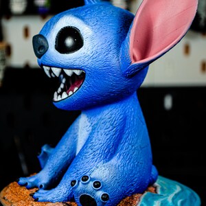 May include: A blue figurine of a cartoon character with large ears, a wide grin, and sharp teeth. The figurine is sitting on a small island with a sandy beach and blue water. The character's fur is textured.