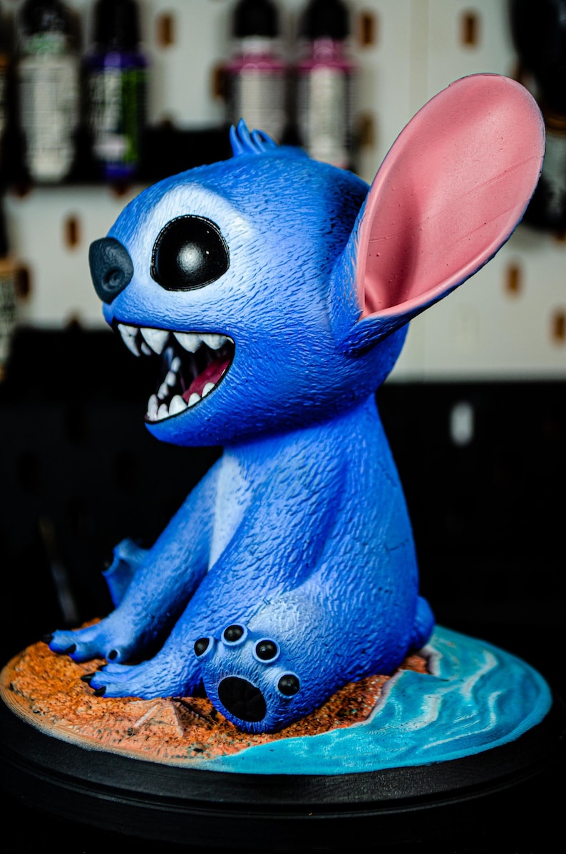 May include: A blue figurine of a cartoon character with large ears, a wide grin, and sharp teeth. The figurine is sitting on a small island with a sandy beach and blue water. The character's fur is textured.