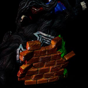 May include: A detailed figurine featuring Spider-Man in a blue and red suit, battling Venom, a black symbiote. A brick wall is breaking apart in the foreground, with green accents. The figures are set against a black background.