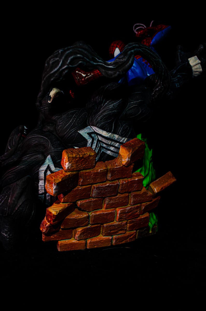 May include: A detailed figurine featuring Spider-Man in a blue and red suit, battling Venom, a black symbiote. A brick wall is breaking apart in the foreground, with green accents. The figures are set against a black background.