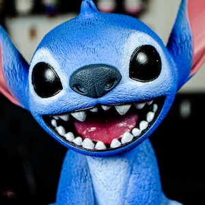 May include: A blue figurine of a cartoon character with large black eyes, a wide grin revealing white teeth, and large ears with pink insides. The character is smiling with its mouth open.