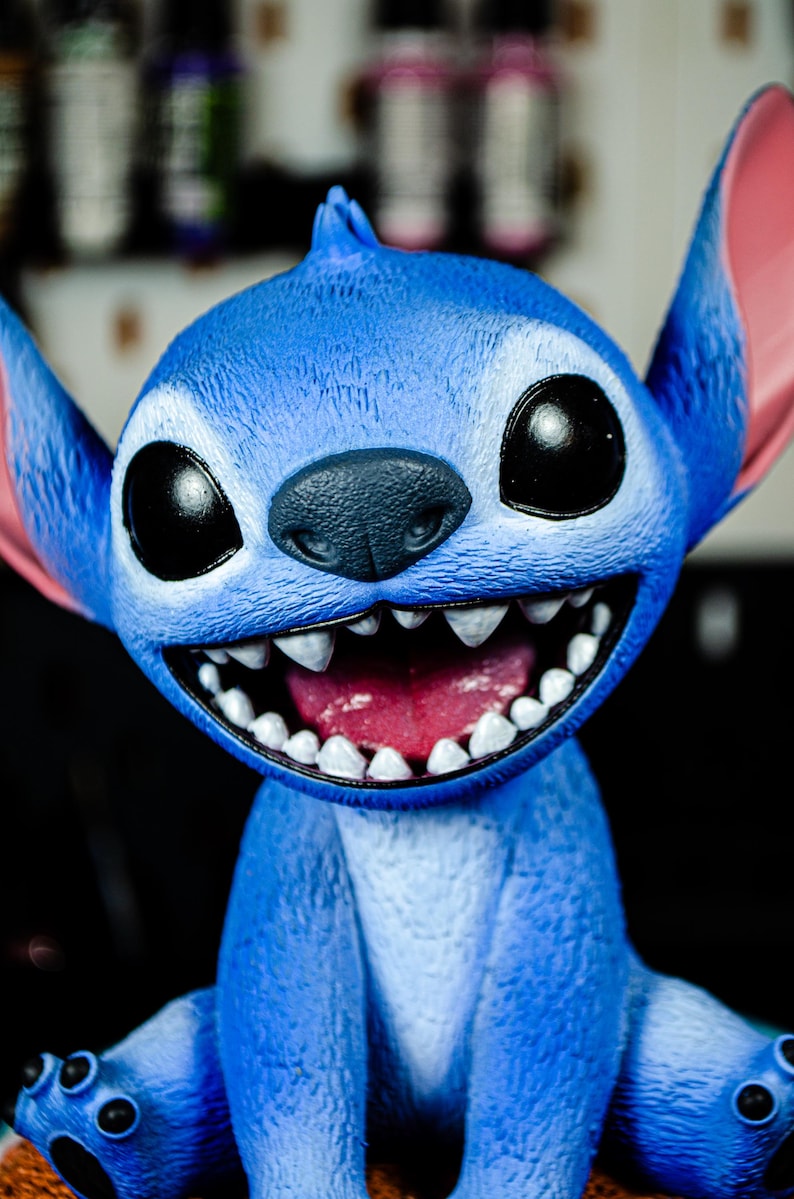 May include: A blue figurine of a cartoon character with large black eyes, a wide grin revealing white teeth, and large ears with pink insides. The character is smiling with its mouth open.