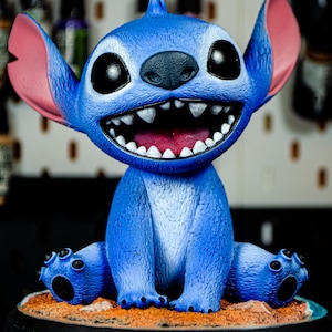 May include: A blue figurine of a cartoon character with large ears, a wide grin, and big eyes. The figurine is sitting on a sandy base with a black circular stand. The character's mouth is open, revealing white teeth and a pink tongue.