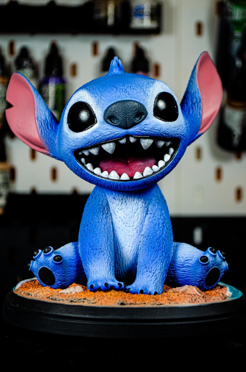 May include: A blue figurine of a cartoon character with large ears, a wide grin, and big eyes. The figurine is sitting on a sandy base with a black circular stand. The character's mouth is open, revealing white teeth and a pink tongue.