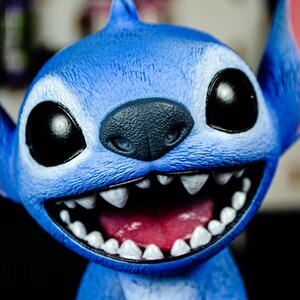 May include: A close-up of a blue character figurine with large black eyes, a dark gray nose, and a wide, toothy grin. The figurine has large ears and a textured surface, with a pink interior mouth.
