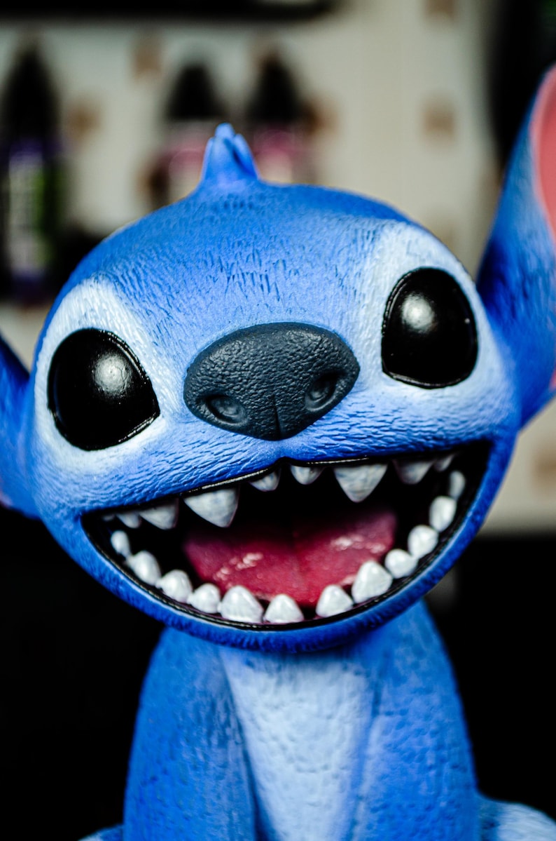 May include: A close-up of a blue character figurine with large black eyes, a dark gray nose, and a wide, toothy grin. The figurine has large ears and a textured surface, with a pink interior mouth.