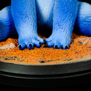 May include: A close-up of a blue figurine's feet and legs. The figurine has textured fur-like skin and black claws. The figurine is standing on a sandy-colored base with a black circular border.