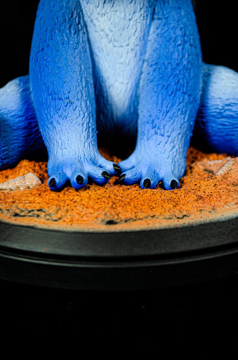 May include: A close-up of a blue figurine's feet and legs. The figurine has textured fur-like skin and black claws. The figurine is standing on a sandy-colored base with a black circular border.