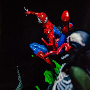 May include: A dynamic action figure of Spider-Man in a classic red and blue suit, mid-leap with web-shooters extended. The figure is set against a dark background, with a green, viscous substance and a blurred figure of Venom.