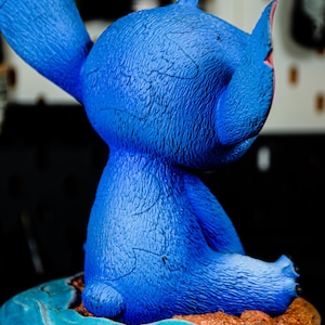 May include: A blue figurine of a cartoon character with large ears and a textured surface. The figurine is seated on a small island with blue water and brown sand. The base is black.