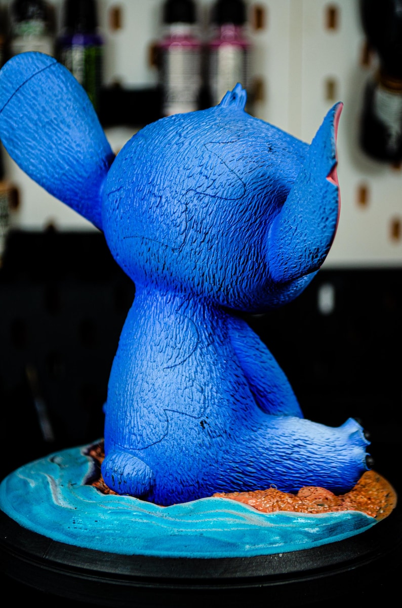 May include: A blue figurine of a cartoon character with large ears and a textured surface. The figurine is seated on a small island with blue water and brown sand. The base is black.