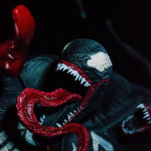 May include: A detailed close-up of a Venom action figure. The figure features a black and white head with sharp white teeth and a long, red tongue. The figure is set against a dark background, highlighting the character's menacing features.