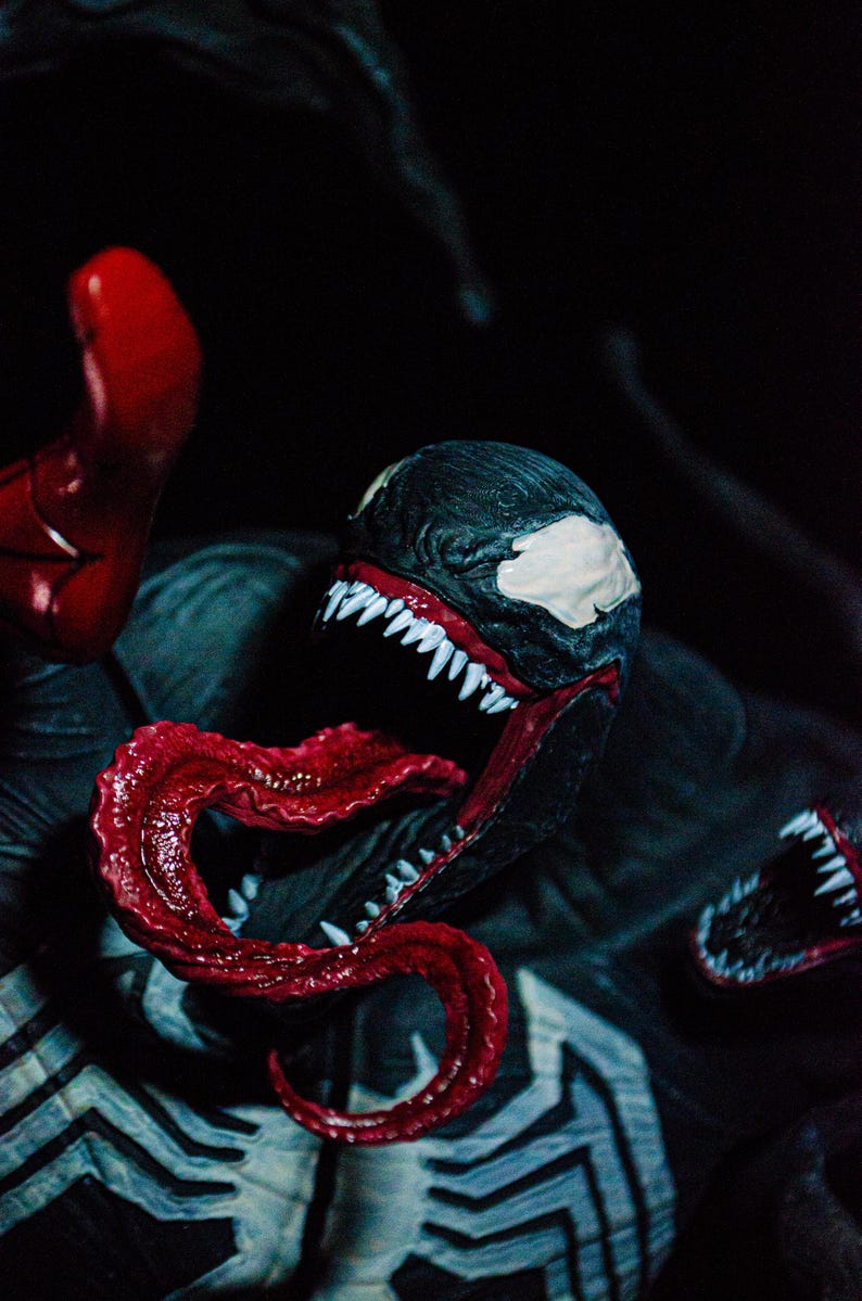 May include: A detailed close-up of a Venom action figure. The figure features a black and white head with sharp white teeth and a long, red tongue. The figure is set against a dark background, highlighting the character's menacing features.