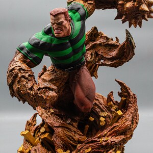 May include: A detailed figurine of a comic book character, Sandman, in a dynamic pose. The figure features a green and black striped shirt, brown skin, and a sand-like texture. Gold coins and bars are at the base.