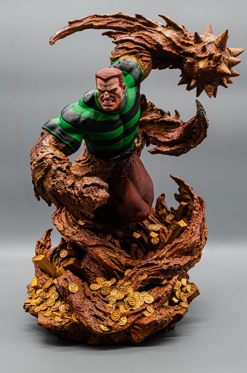 May include: A detailed figurine of a comic book character, Sandman, in a dynamic pose. The figure features a green and black striped shirt, brown skin, and a menacing expression. The base is adorned with gold coins and a textured, brown, sand-like material.