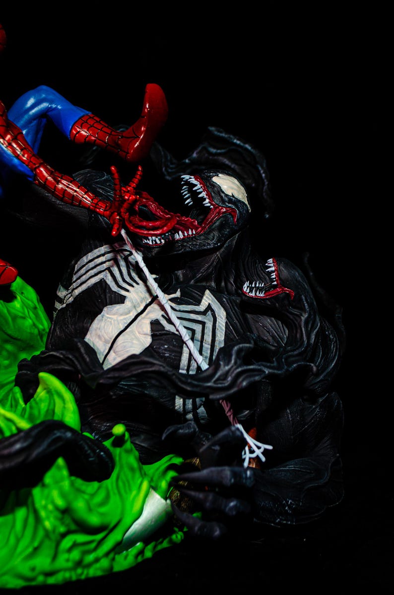 May include: A detailed figurine of Venom in a battle scene with Spider-Man. Venom is predominantly black with white accents and a large, menacing mouth. Spider-Man is in a classic red and blue suit, with his web-shooters visible.