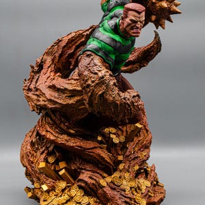 May include: A detailed statue of a comic book character, Sandman, in a dynamic pose. The figure is predominantly brown and tan, with a green and black striped shirt. The base features gold coins and bars, with a gray background.