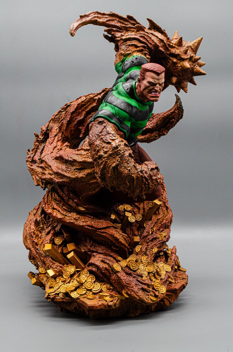 May include: A detailed statue of a comic book character, Sandman, in a dynamic pose. The figure is predominantly brown and tan, with a green and black striped shirt. The base features gold coins and bars, with a gray background.