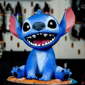 May include: A blue figurine of the character Stitch from Lilo & Stitch. The figurine has large black eyes, a wide grin with visible teeth, and large ears. It is sitting on a sandy base with a black border.