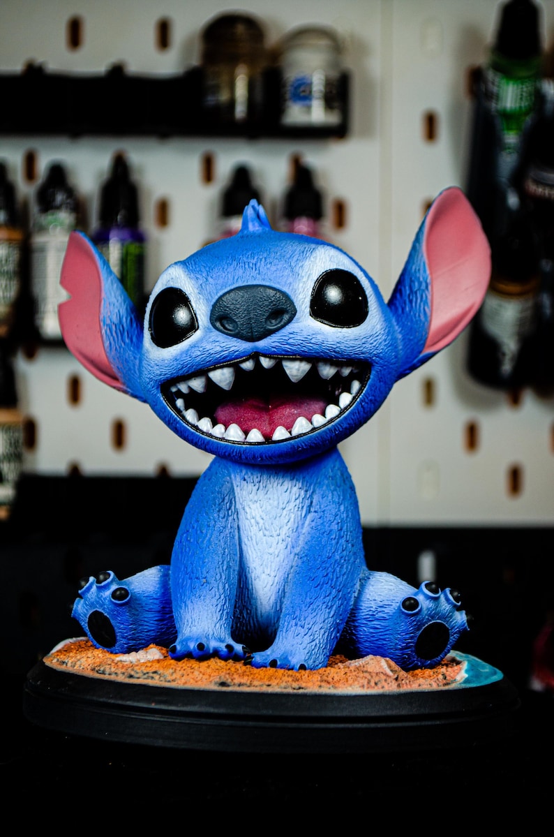 May include: A blue figurine of the character Stitch from Lilo & Stitch. The figurine has large black eyes, a wide grin with visible teeth, and large ears. It is sitting on a sandy base with a black border.