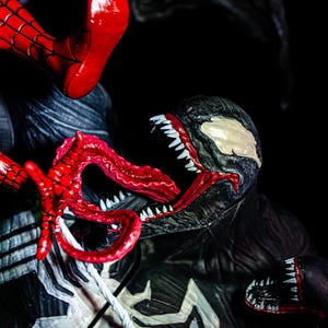 May include: A detailed action figure scene featuring Spider-Man and Venom. Spider-Man's red and blue costume is visible, along with Venom's black and white design, open mouth, and sharp teeth. The figures are set against a dark background.