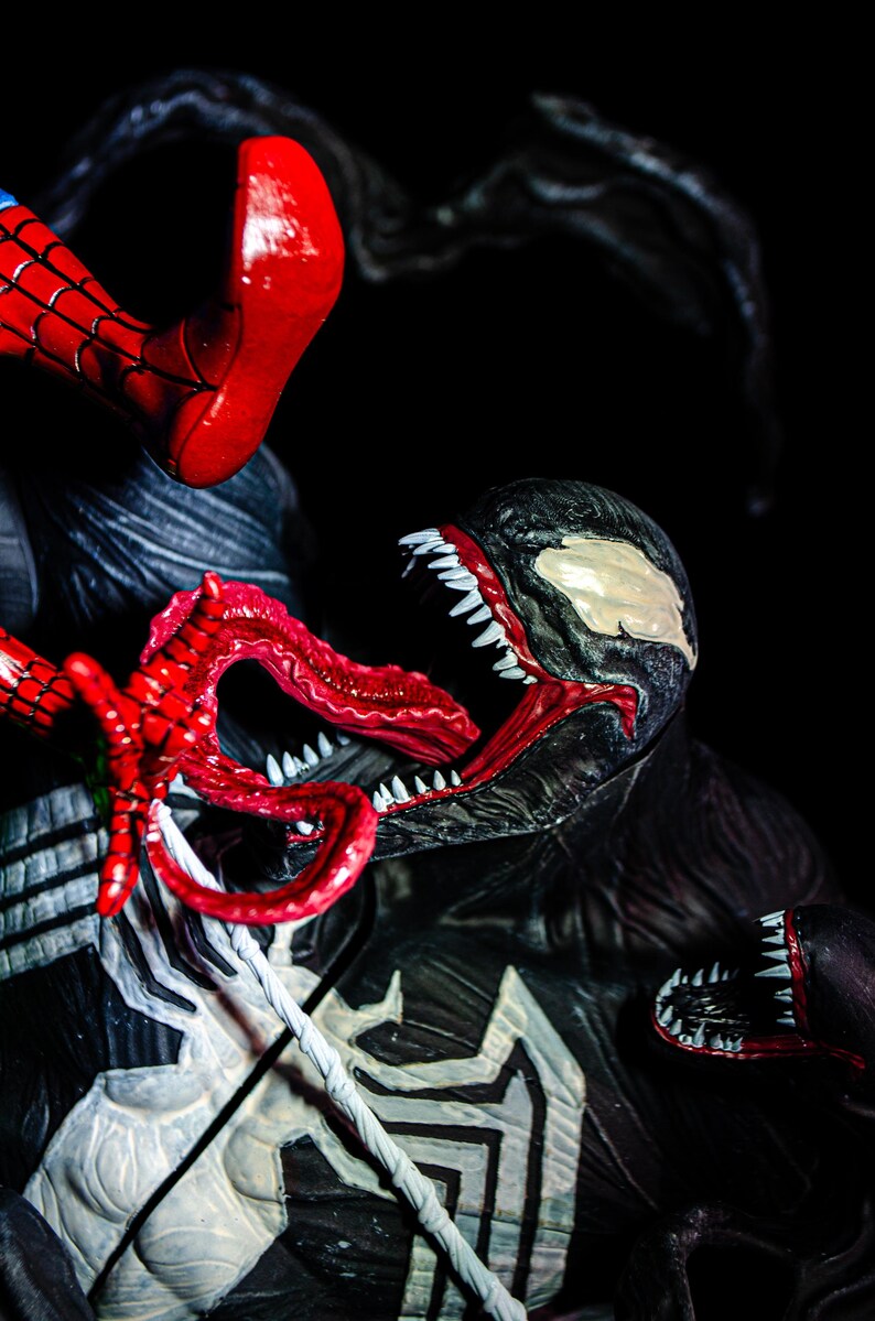 May include: A detailed action figure scene featuring Spider-Man and Venom. Spider-Man's red and blue costume is visible, along with Venom's black and white design, open mouth, and sharp teeth. The figures are set against a dark background.