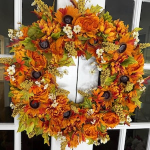 May include: A vibrant autumn wreath featuring sunflowers, orange roses, and various fall foliage. The wreath is adorned with small white flowers and yellow accents, creating a warm and inviting seasonal decoration.