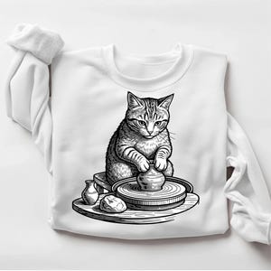 May include: White sweatshirt featuring a black and white illustration of a cat crafting pottery on a pottery wheel. The cat is seated on a small stool, with a focused expression. Additional pottery items are on a wooden surface.