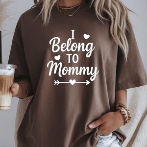 May include: A brown t-shirt with the white text "I Belong to Mommy". The text includes a heart symbol above the word "Belong" and a heart symbol above the word "Mommy". The shirt has short sleeves.