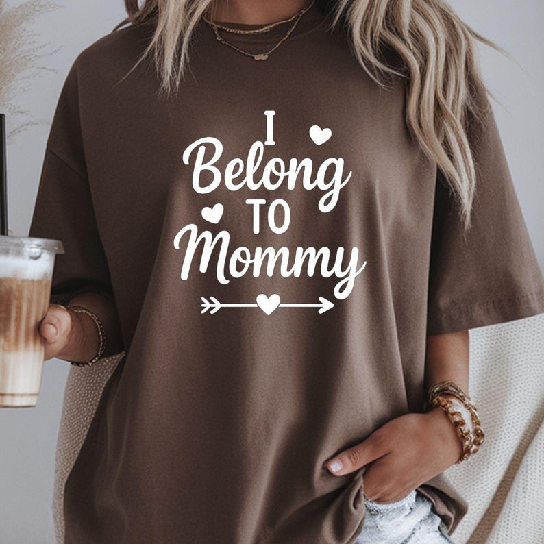 May include: A brown t-shirt with the white text "I Belong to Mommy". The text includes a heart symbol above the word "Belong" and a heart symbol above the word "Mommy". The shirt has short sleeves.