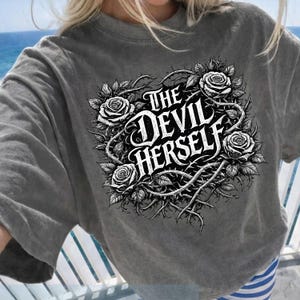 Puede incluir: Una camiseta gris de manga corta con el texto "THE DEVIL HERSELF" en una fuente gótica, rodeada de rosas y espinas. La camiseta tiene un aspecto vintage y desgastado.