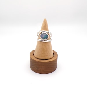 May include: A silver ring with an oval, iridescent stone is displayed on a wooden ring holder. The ring has a double-band design, with the stone set in the center. The ring holder is a light brown cone shape, set on a dark brown, round base.