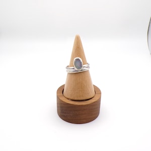 May include: Two silver rings with a light blue oval stone are displayed on a wooden ring holder. The rings are stacked, showcasing the design and texture of the metal and stone. The holder is a natural wood color.
