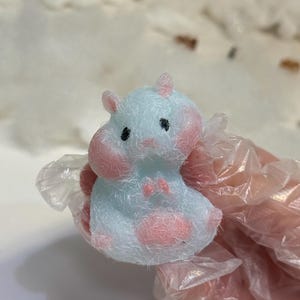May include: A light blue hippo figurine with pink accents on the cheeks, ears, and belly. The hippo has small black eyes and a fuzzy texture. The figurine is held in a clear plastic glove.