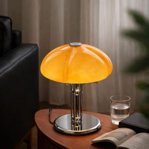 Puede incluir: Una lámpara de mesa de estilo retro con base y tallo cromados, coronada por una pantalla en forma de cúpula de color naranja-amarillo vibrante. La lámpara está sobre una mesa de madera, junto a un vaso de agua y un libro abierto.