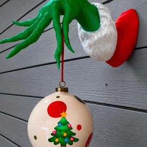 May include: A wall-mounted green hand with long fingers emerging from a red sleeve with a white faux fur cuff. The hand holds a red string attached to a round white Christmas ornament decorated with a green tree graphic and red dots.